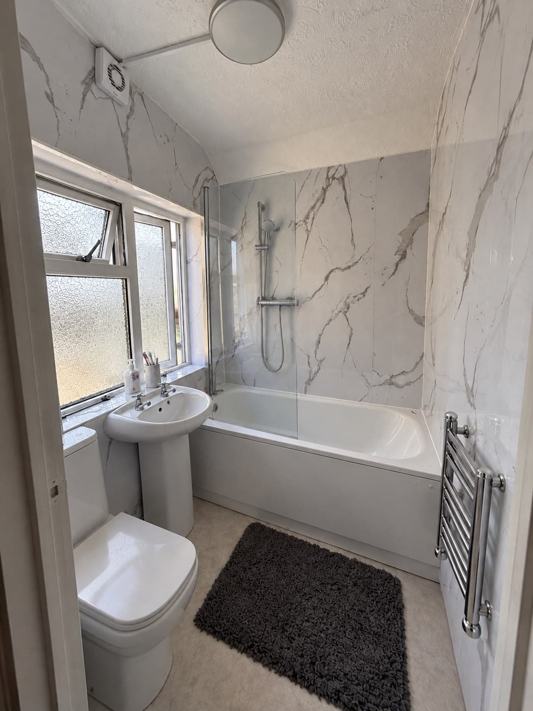 Upgraded family bathroom with marble-effect wall panels, bath with glass shower screen, pedestal basin, WC and chrome towel radiator